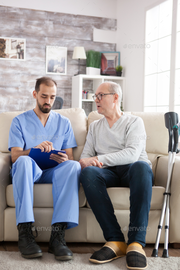 Male doctor with senior man sitting on couch in nursing home writing notes on clipboard