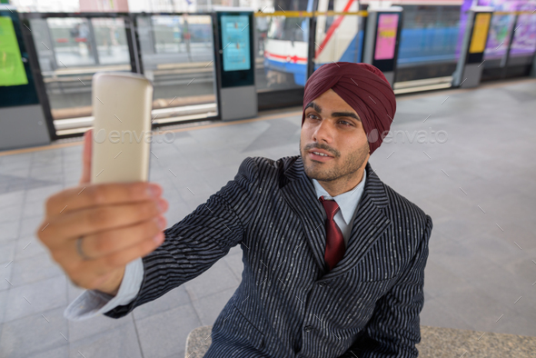 Indian businessman sitting at train station while taking selfie with mobile phone