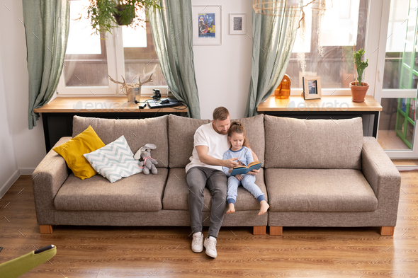 Young father and his cute little daughter sitting on couch and reading book