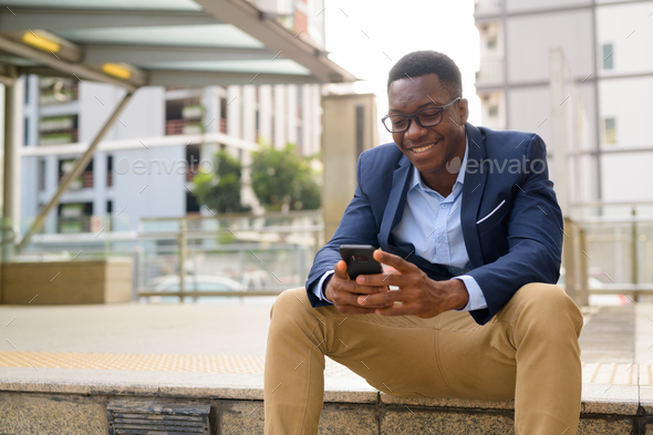 Young happy African businessman using phone while sitting outside the subway train station