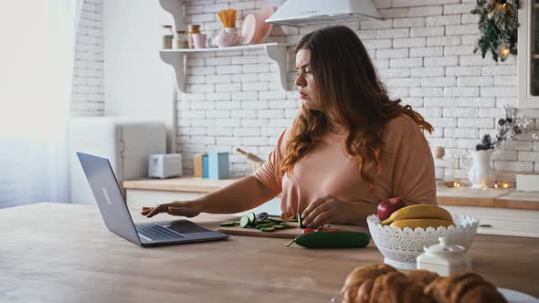 Overweight Lady Cutting Cucumber at Kitchen and Watching Soap Opera Show on Laptop alt