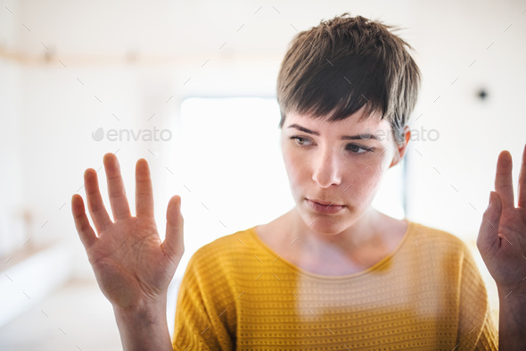 Front view of sad young woman standing indoors at home, hands on glass.