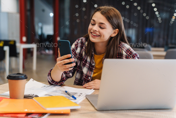 Image of smiling girl using cellphone while doing homework with laptop