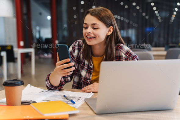 Image of smiling girl using cellphone while doing homework with laptop