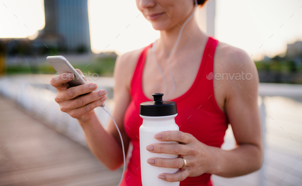 Midsection of young woman runner with earphones in city, using smartphone
