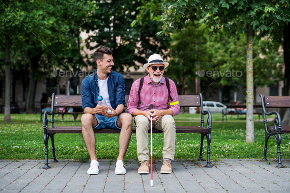 Young man and blind senior with white cane sitting on bench in park in city