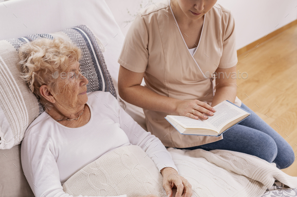 Helpful young carer reads a book to an elderly woman lying in a hospital bed