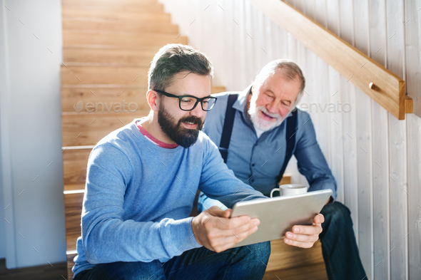 An adult son and senior father with tablet sitting on stairs indoors at home