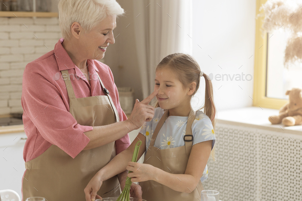 Fun baking time. Happy older lady touching her granddaughter\'s nose in kitchen, empty space