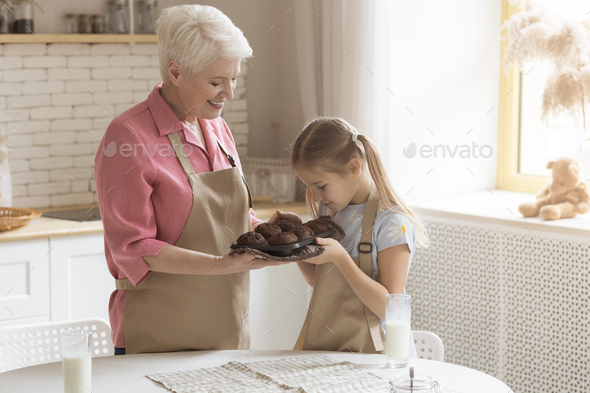 Little girl enjoying aroma of fresh chocolate muffins with granny in kitchen, blank space