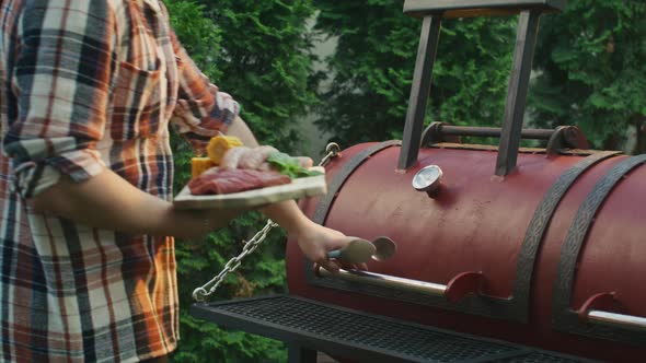 Man with a Plate of Meat and Sausages Approaches the Grill and Begins Cooking