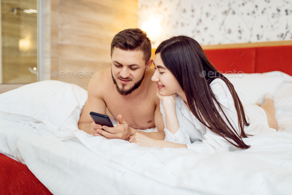 Love couple in pajamas using phone in bed at home