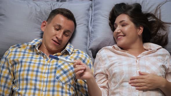 Young Woman Shows a Man a Pregnancy Test While Lying in Bed Reverie Top View alt