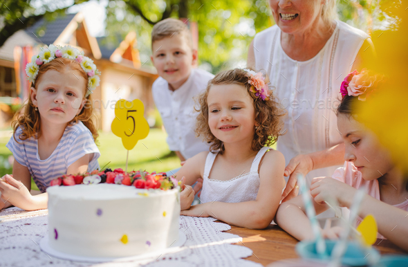 Children with cake standing around table on birthday party in garden in summer.