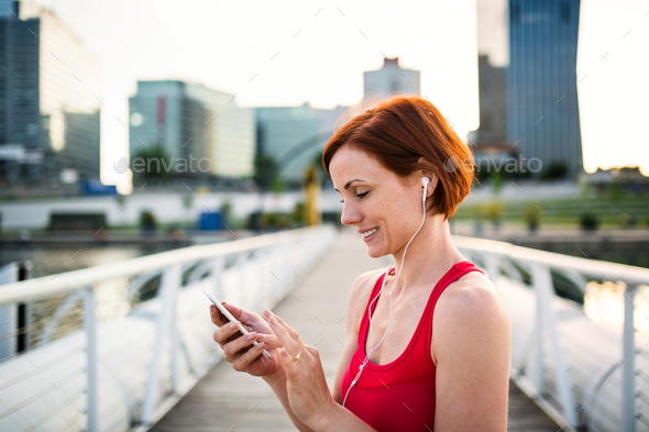 Young woman runner with earphones in city, using smartphone