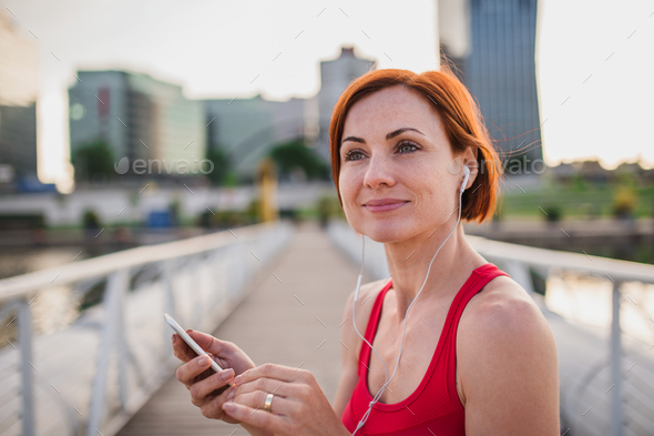 Young woman runner with earphones in city, using smartphone