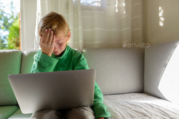 Stressed young boy using laptop and getting bad news on the couch at home