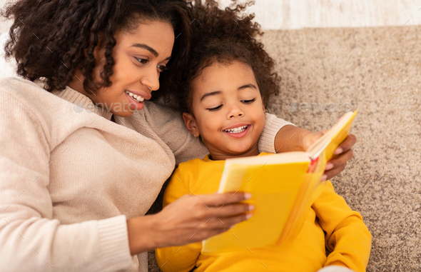 Cute Mother And Little Daughter Reading Book Sitting On Couch