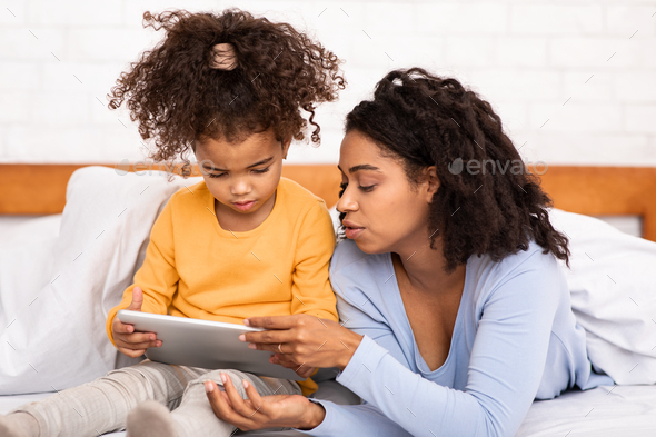 Mother Teaching Daughter To Use Tablet Lying In Bed Indoors