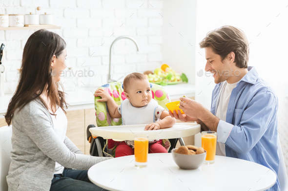 Young parents feeding baby son with breakfast at kitchen