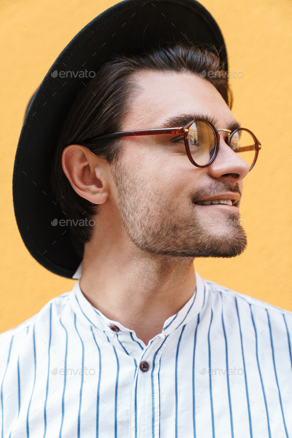 Image of young cheerful man wearing eyeglasses and black hat smiling and looking aside at copyspace
