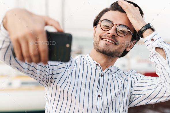 Photo of happy brunette man taking selfie photo on smartphone and smiling while walking on pier