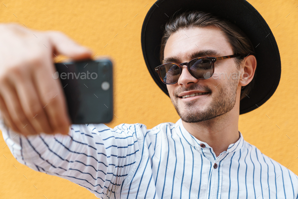 Image of young cheerful man wearing sunglasses and black hat smiling and taking selfie at cellphone