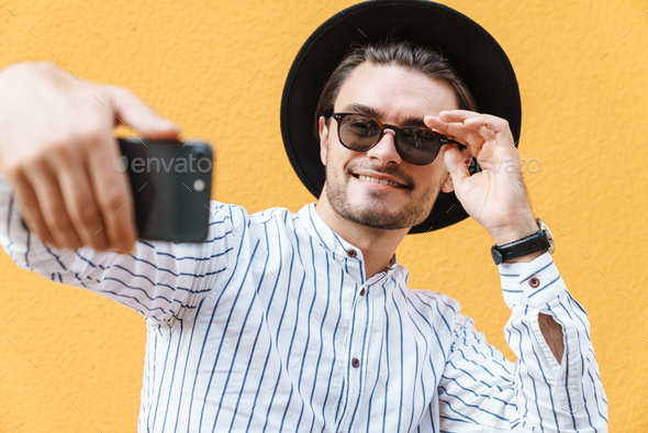 Image of young joyful man wearing sunglasses and black hat smiling and taking selfie at cellphone