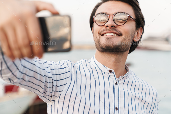 Photo of cheerful caucasian man taking selfie photo on smartphone and smiling while walking on pier