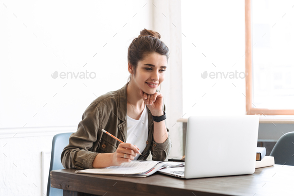 Woman sitting at desk, using computer and writing in notebook