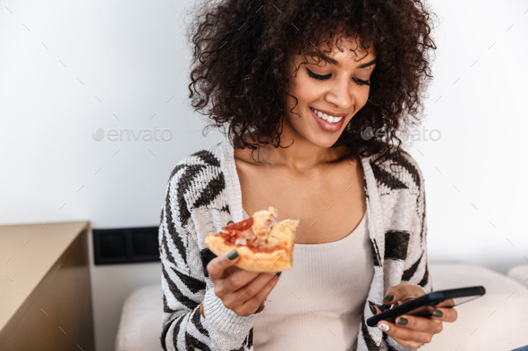 Pretty young african woman indoors at home eat pizza.