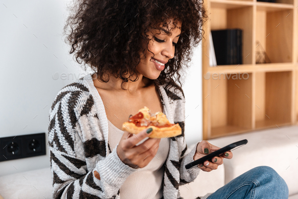 Pretty young african woman indoors at home eat pizza.