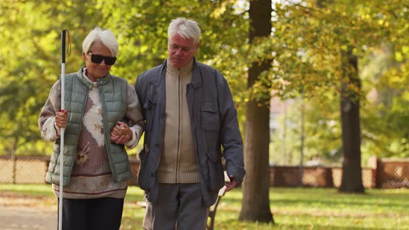 Elderly Caucasian Man Supports His Blind Wife While Walking in the Park Disabled Blind People alt