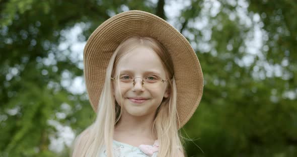 Cute Little Girl Model with Glasses and Hat Smiling and Looks in Camera in Park alt