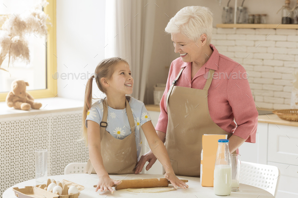 Pizza time. Cute girl with her granny rolling dough for pastry together in kitchen, empty space