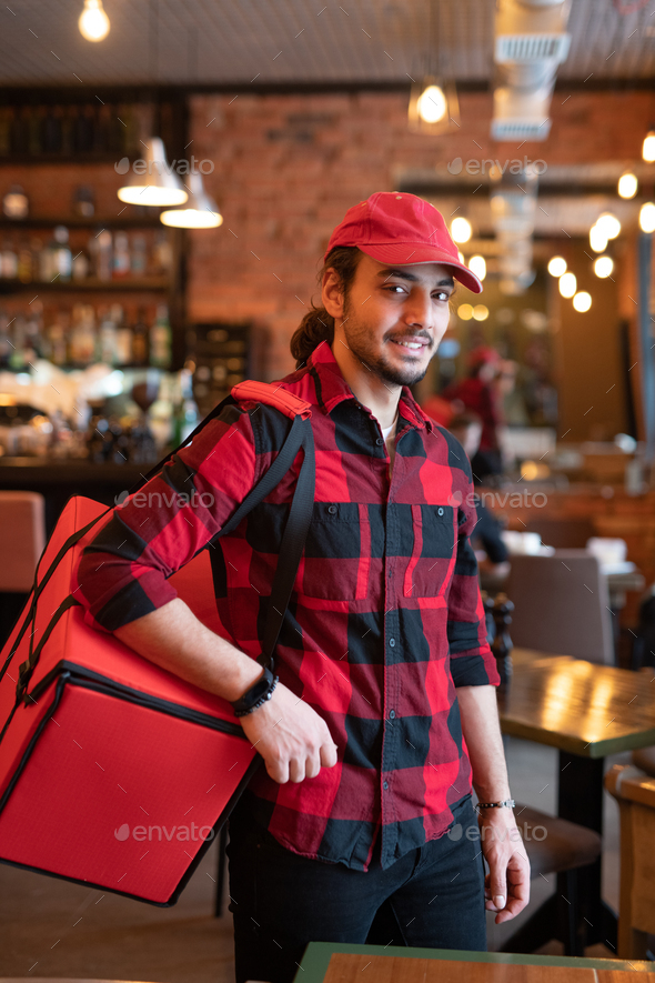 Young courier in red cap and flannel carrying big bag on shoulder