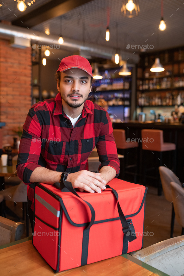 Young handsome courier in red flannel and cap standing by big bag with orders
