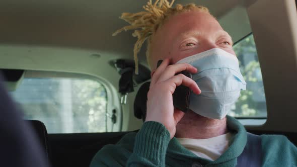 Happy albino african american man wearing face mask sitting in car talking on smartphone alt