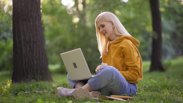 Young Blonde Woman Happily Talking By Video Call Using Laptop on Green Grass alt