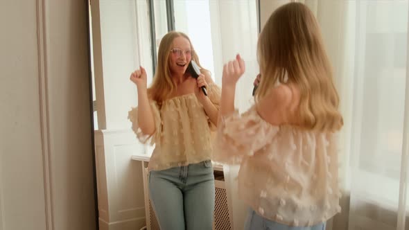 A young woman sings into a hairbrush as like microphone in front of a mirror dance alt