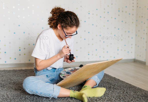 Curly hair girl teenager in glasses in white t-shirt painting on wooden desk