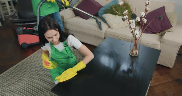 Woman-Cleaner in Apron and Gloves which Wiping Wooden Table with Detergent alt