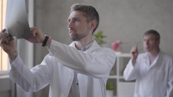 Two Male Doctors Checking X-ray in the Office of Modern Clinic. Younger Man in White Robe Showing x alt