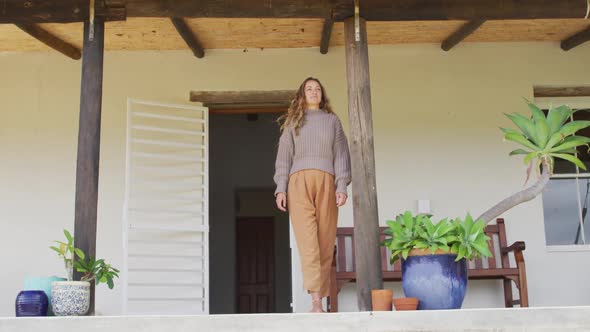 Smiling caucasian woman standing, leaning outside house looking out from terrace alt