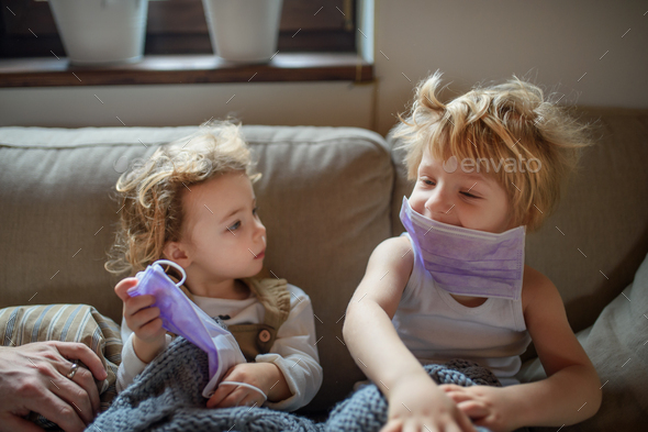 Two small sick children with face mask at home sitting on sofa
