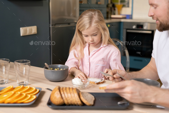 Pretty blond little girl spreading butter on slice of wheat bread by breakfast
