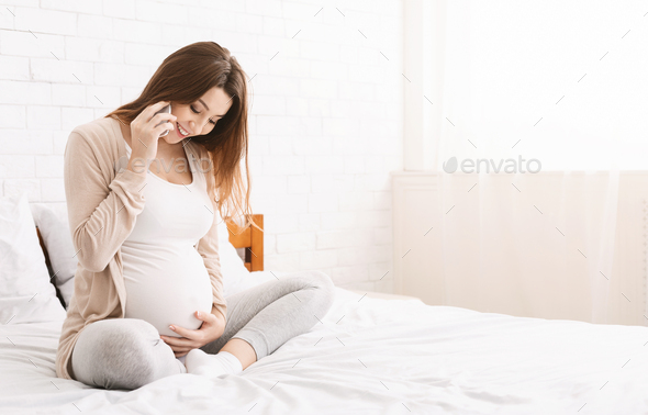 Pregnant woman talking on phone and embracing her belly at home