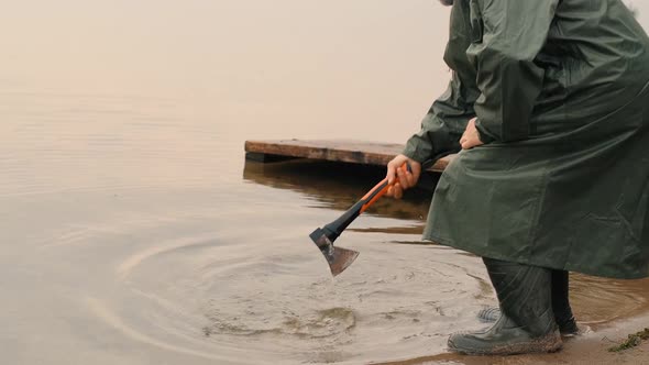 An Elderly Male Tourist in a Raincoat Rinses an Axe in a Pond on an Autumn Evening at a Halt