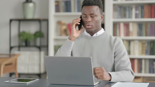 African Man Talking on Smartphone While Using Laptop in Library alt