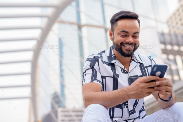 Happy young bearded Indian man using phone and sitting in the city outdoors - Stock Photo - Images